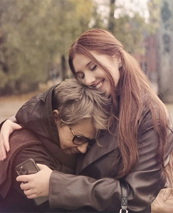 A young woman with long reddish-brown hair smiling while hugging an older woman with short grey hair in an outdoor park.