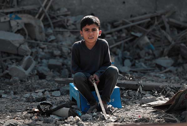A young boy sitting on a blue crate amidst the rubble of destroyed buildings, highlighting global social issues and the need for cross-cultural understanding.